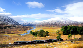 Scottish Winter train This is a landscape photograph taken in the late morning during winter in Scotland, showcasing a Scottish Winter train traveling through the rural Highlands. The scene highlights the natural beauty of the United Kingdom, featuring mountains partially covered with snow, a meandering river running through the valley, and stretches of evergreen trees. The train, serving as the main subject and representing a vital mode of transport in this remote region, moves through a picturesque winter landscape surrounded by nature and rugged Scottish terrain. The snow-dusted peaks and the flowing water emphasize the season, and demonstrate the connection between transport infrastructure and the rural environment in Scotland.